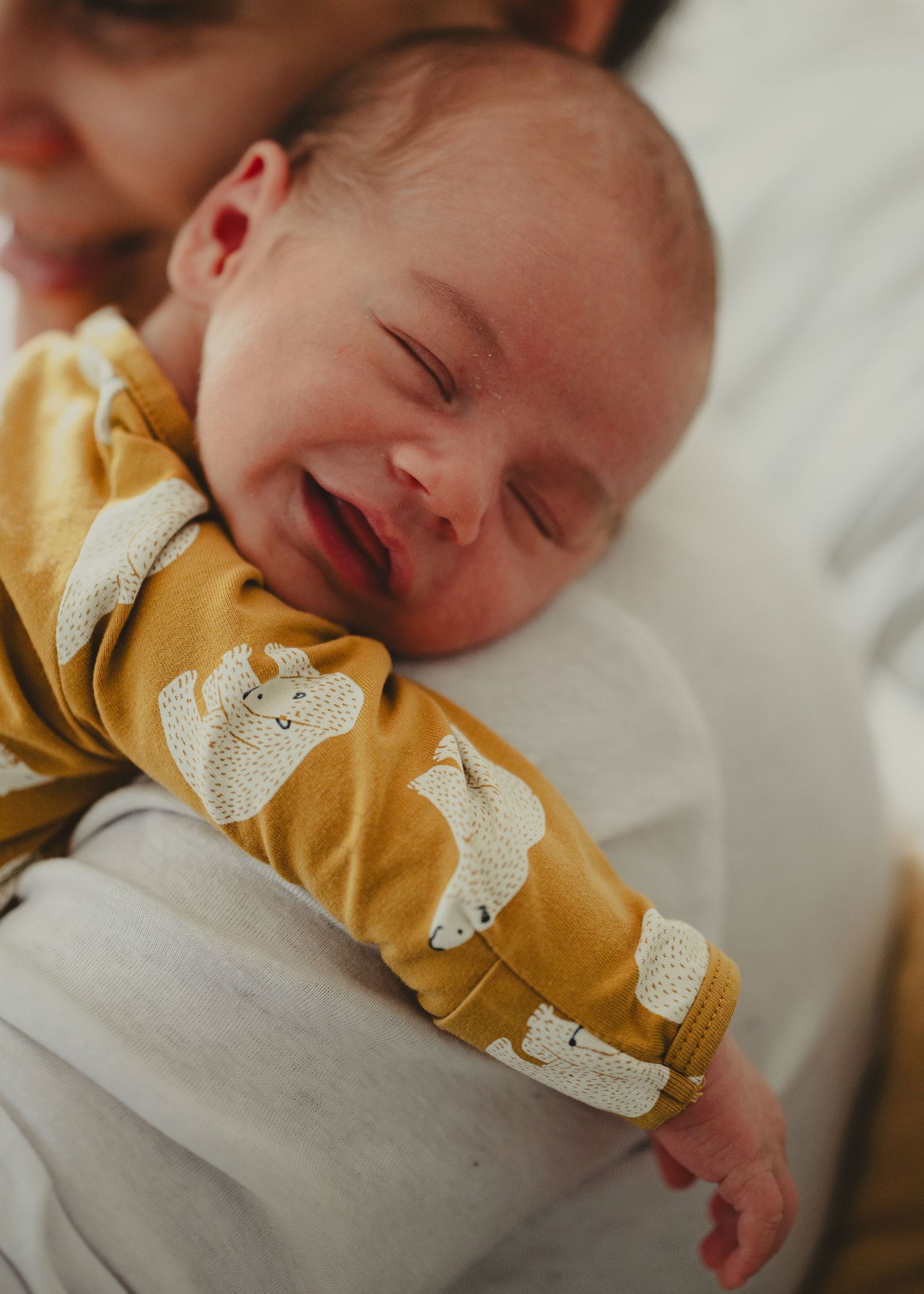 Smiling baby in yellow onesie with cute bear print lying down on white bedding.