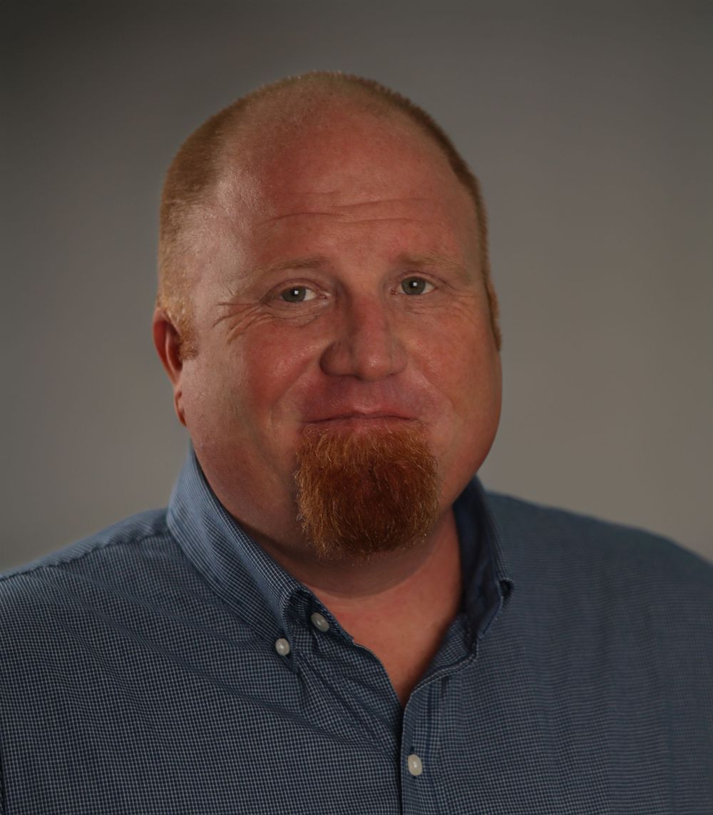 Professional headshot portrait of person in blue button-down collar shirt against gray backdrop.
