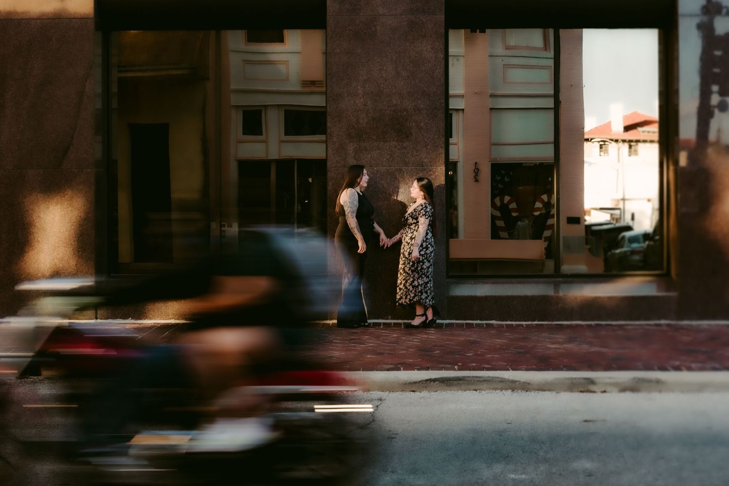 A couple stands on a street at night while a blurred motorbike passes in front of them, with warm storefront lighting.