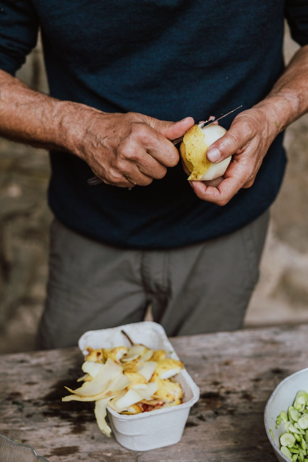 Hands peeling a lemon over a container of sliced ingredients on a rustic wooden surface.