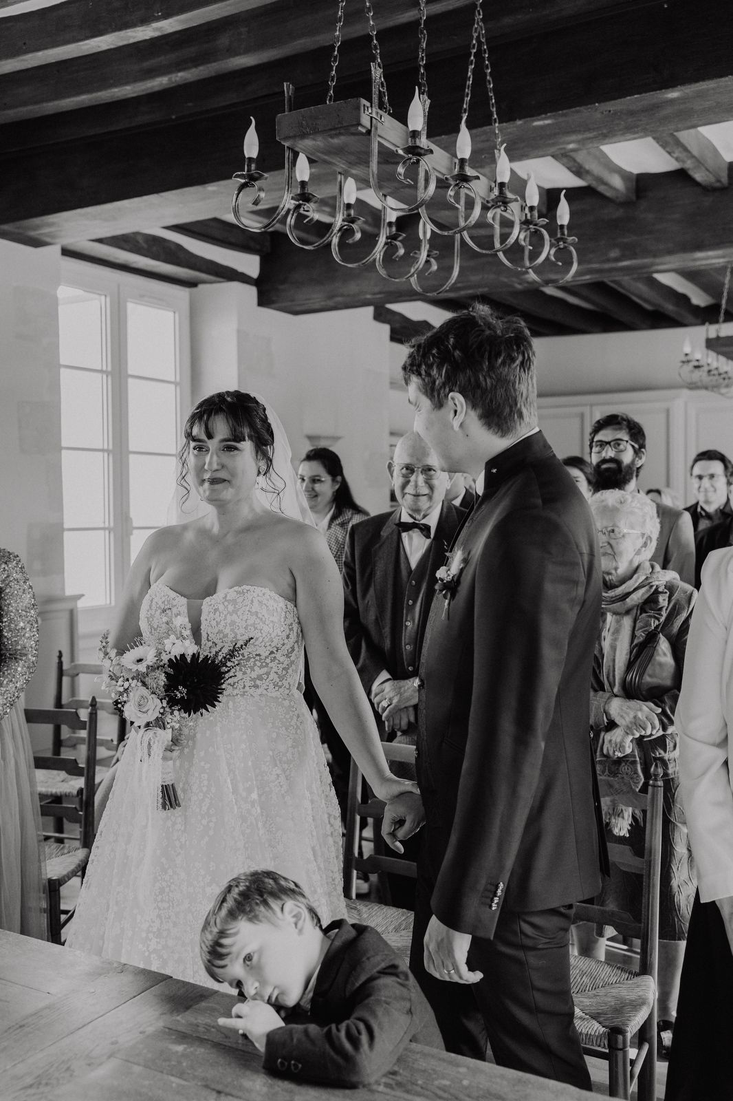 Wedding ceremony moments captured in black and white showing guests and a chandelier in an indoor venue.