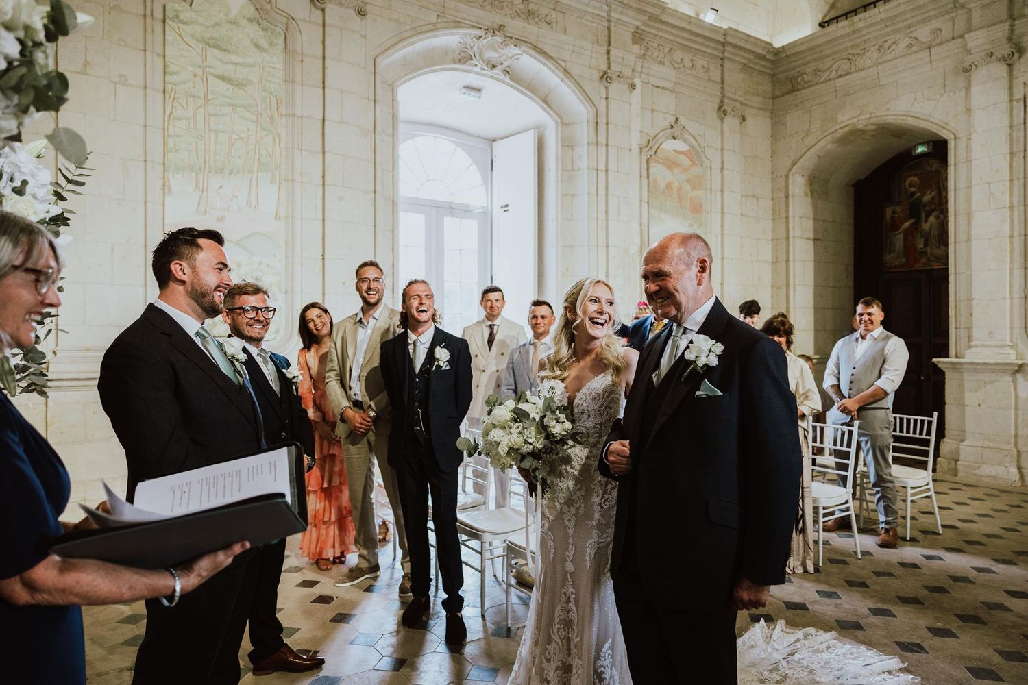 Group of elegantly dressed guests gather in conversation during a wedding celebration in a historic stone building interior.