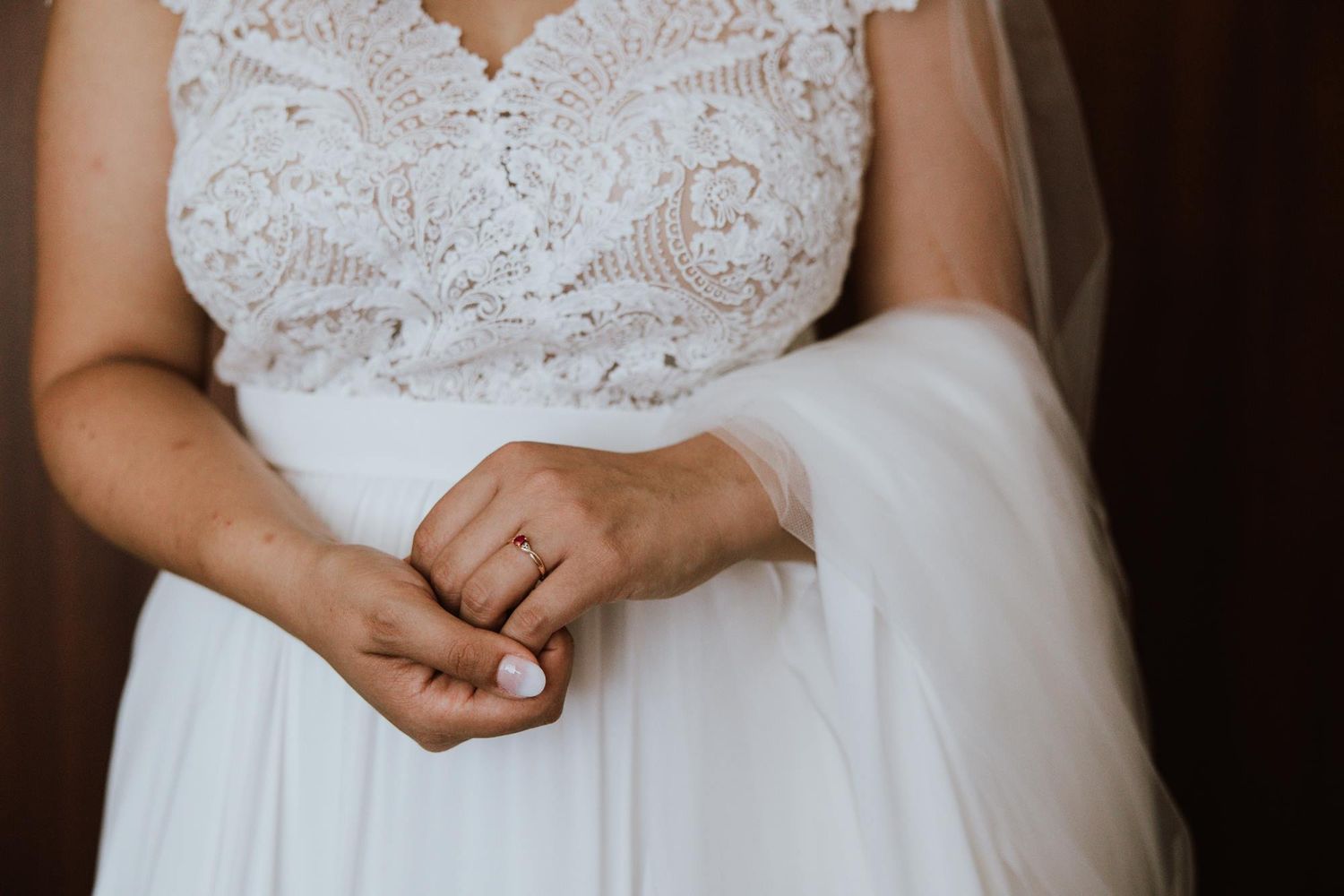 Close up of a wedding dress showing delicate white lace bodice detail and flowing tulle skirt with hands clasped in front.