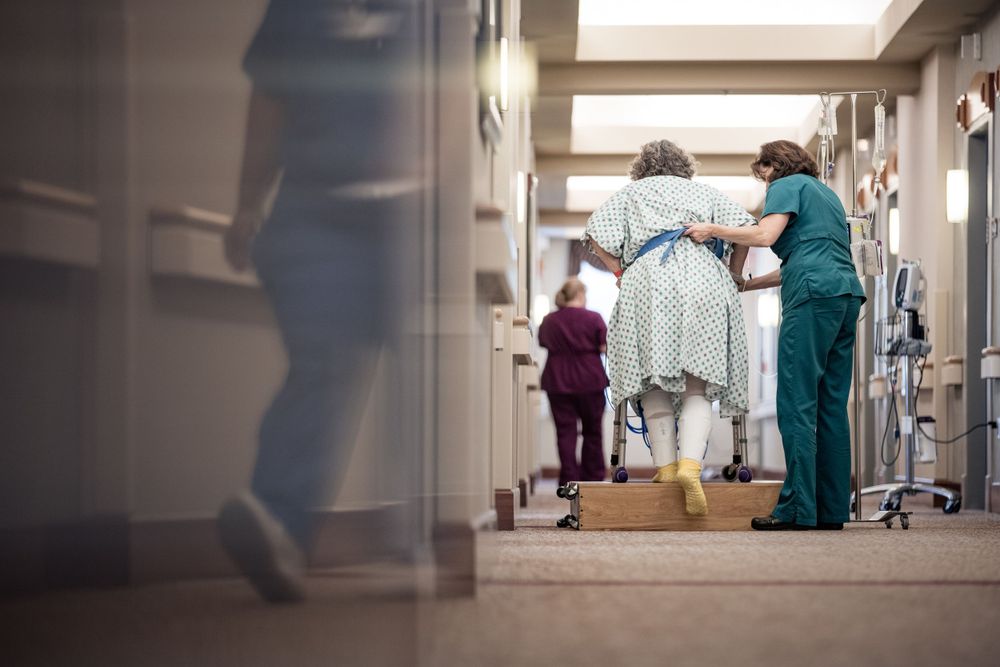 Healthcare worker assists patient walking in hospital hallway while using mobility assistance device.