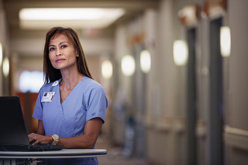 Healthcare worker in blue scrubs works at a hospital computer station in a softly lit corridor.