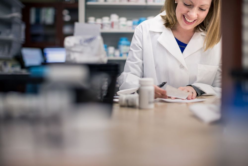 Healthcare professional in white lab coat writing notes at pharmacy counter with medical supplies in background.