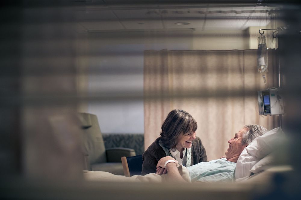Hospital scene showing patient in bed receiving care while visitor sits nearby in dimly lit room.
