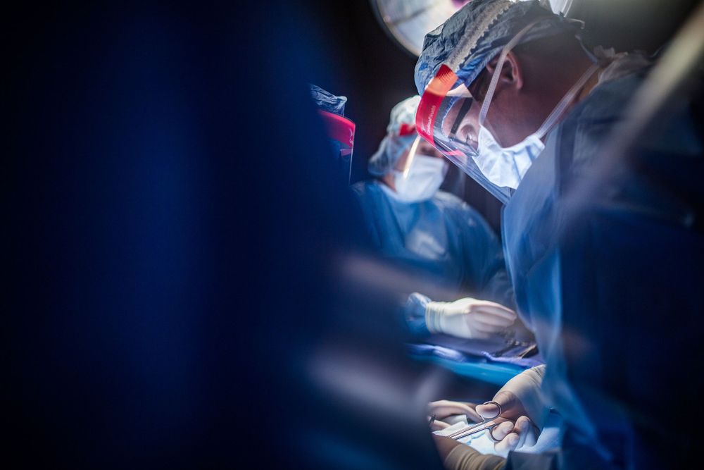 Medical team performs surgery in an operating room with blue surgical drapes and bright lighting.