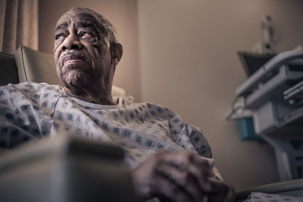 Elderly patient in hospital gown rests in medical setting with equipment visible in soft lighting.