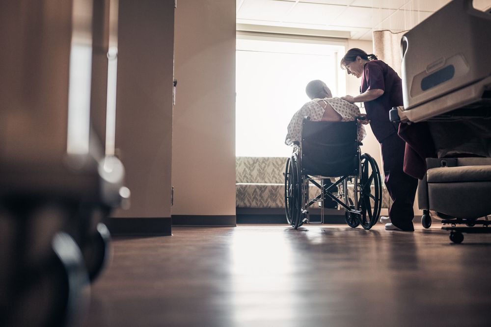 A healthcare worker assists a patient in a wheelchair in a dimly lit hospital hallway.
