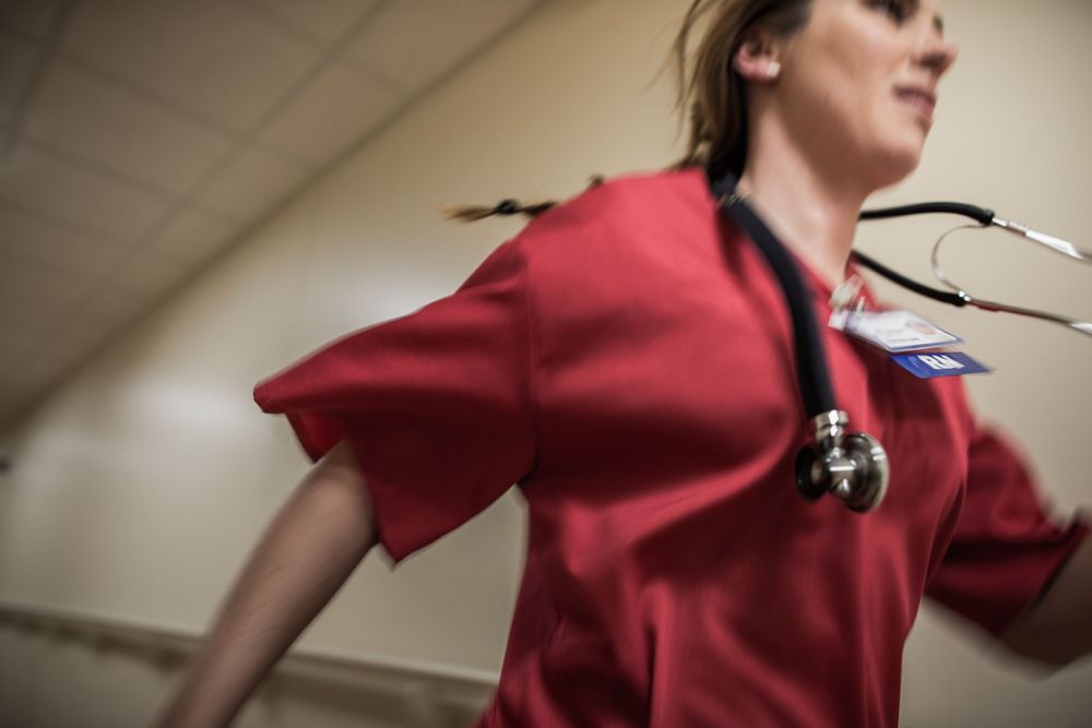 A medical professional in red scrubs walks through a hospital hallway wearing a stethoscope.