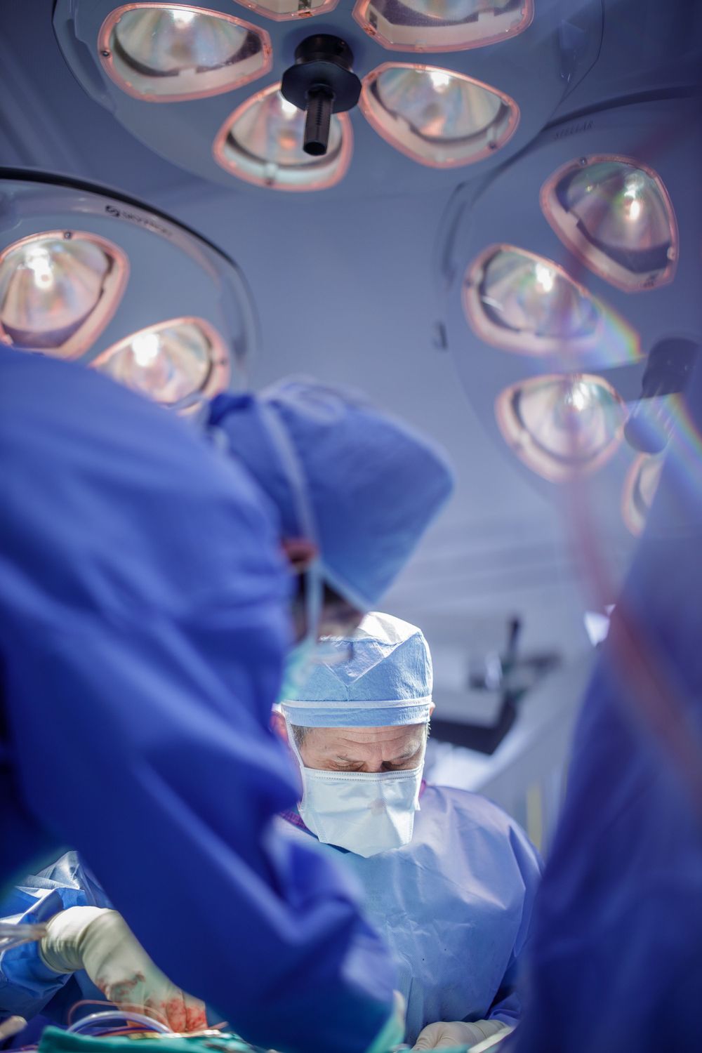 Medical team wearing blue scrubs performs surgery under bright operating room lights.