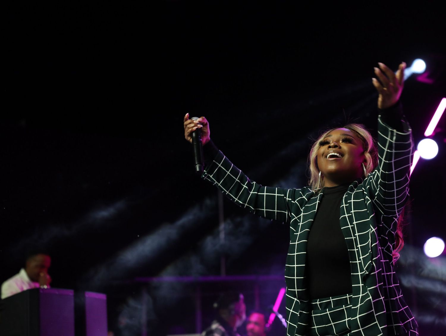 Performer in black and white checkered outfit singing passionately on stage with arms raised under purple lighting.