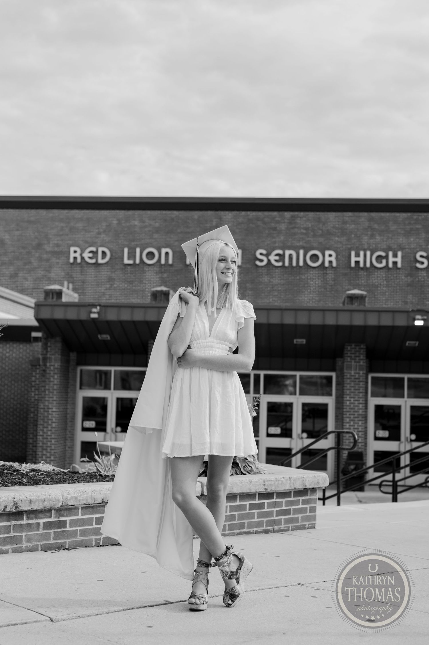 Black and white photo of graduate posing by school entrance in flowing cape.