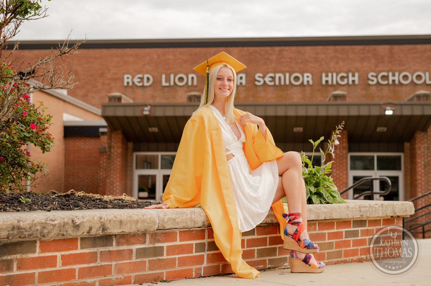 Graduate in yellow cap and gown sits casually on brick ledge at school entrance.