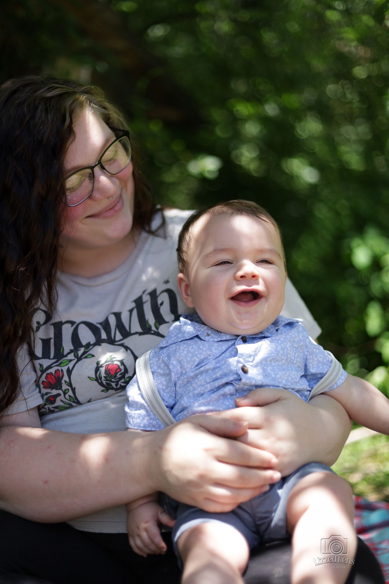 A laughing baby boy sits on his mother's lap outdoors in a sunny green garden.