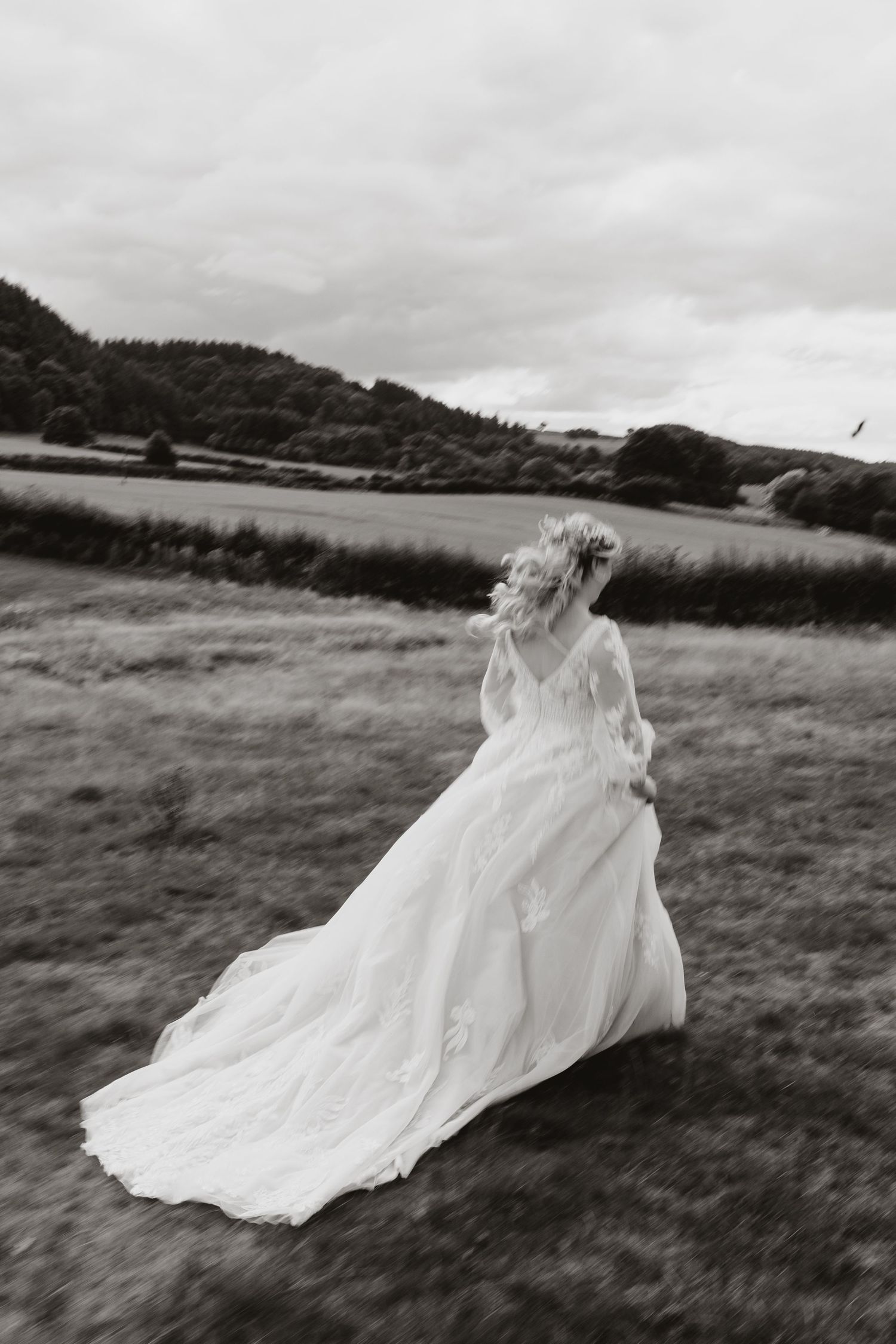 A flowing white wedding gown billows dramatically in the wind across a grassy field with hills in the background.