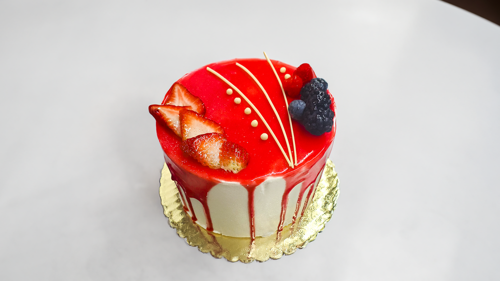 Small round cake with red glaze, strawberry slices, blueberry, blackberry, white chocolate drizzles on gold doily.