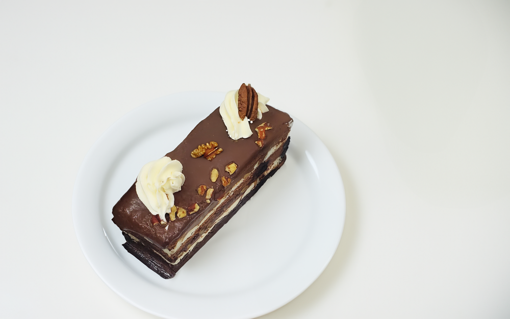 Slice of layered chocolate cake bar with frosting, cream dollop, and pecans on a white plate.