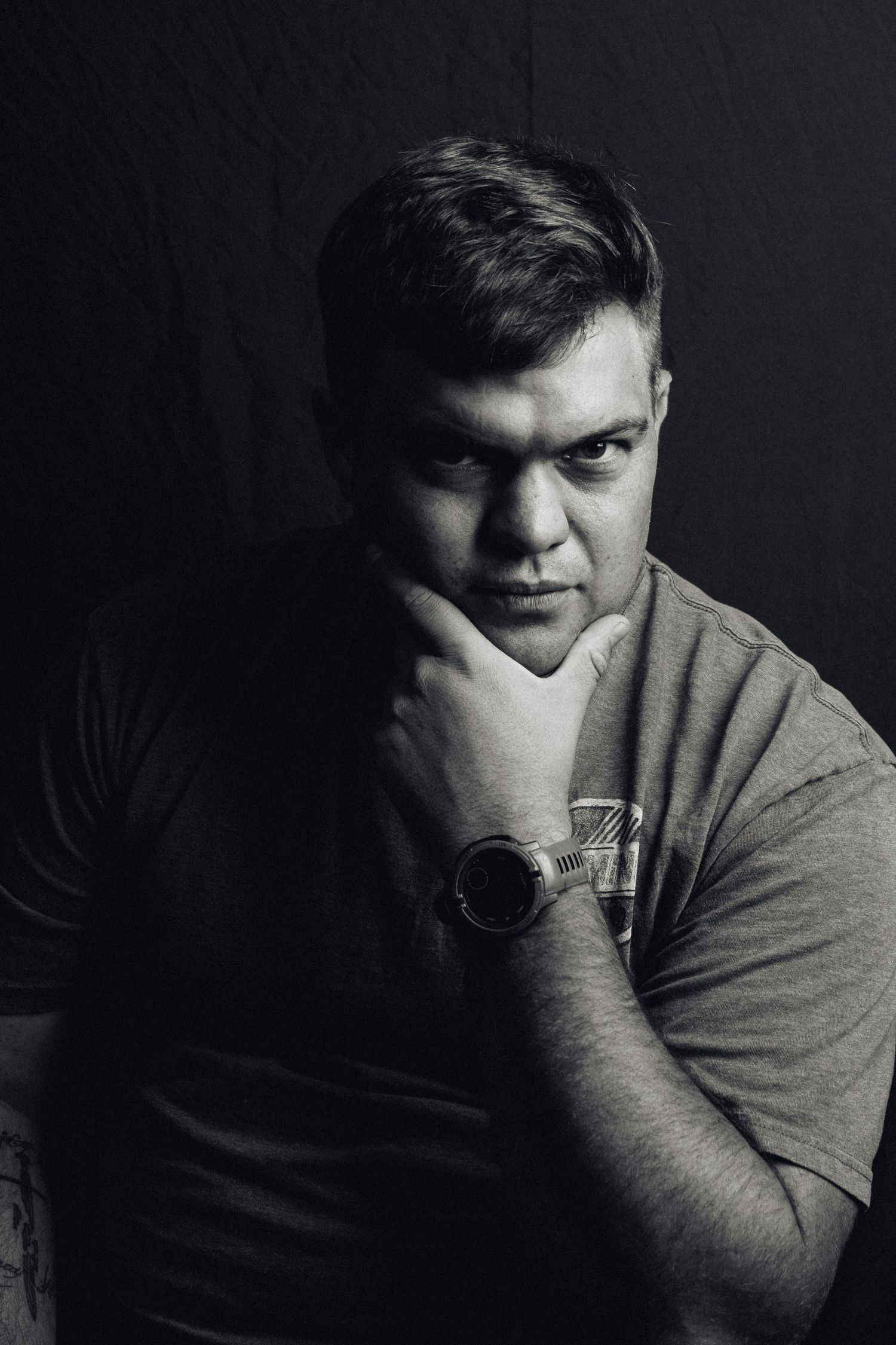 A dramatic black and white portrait shows a contemplative figure in a t-shirt with hand on chin against a dark background.