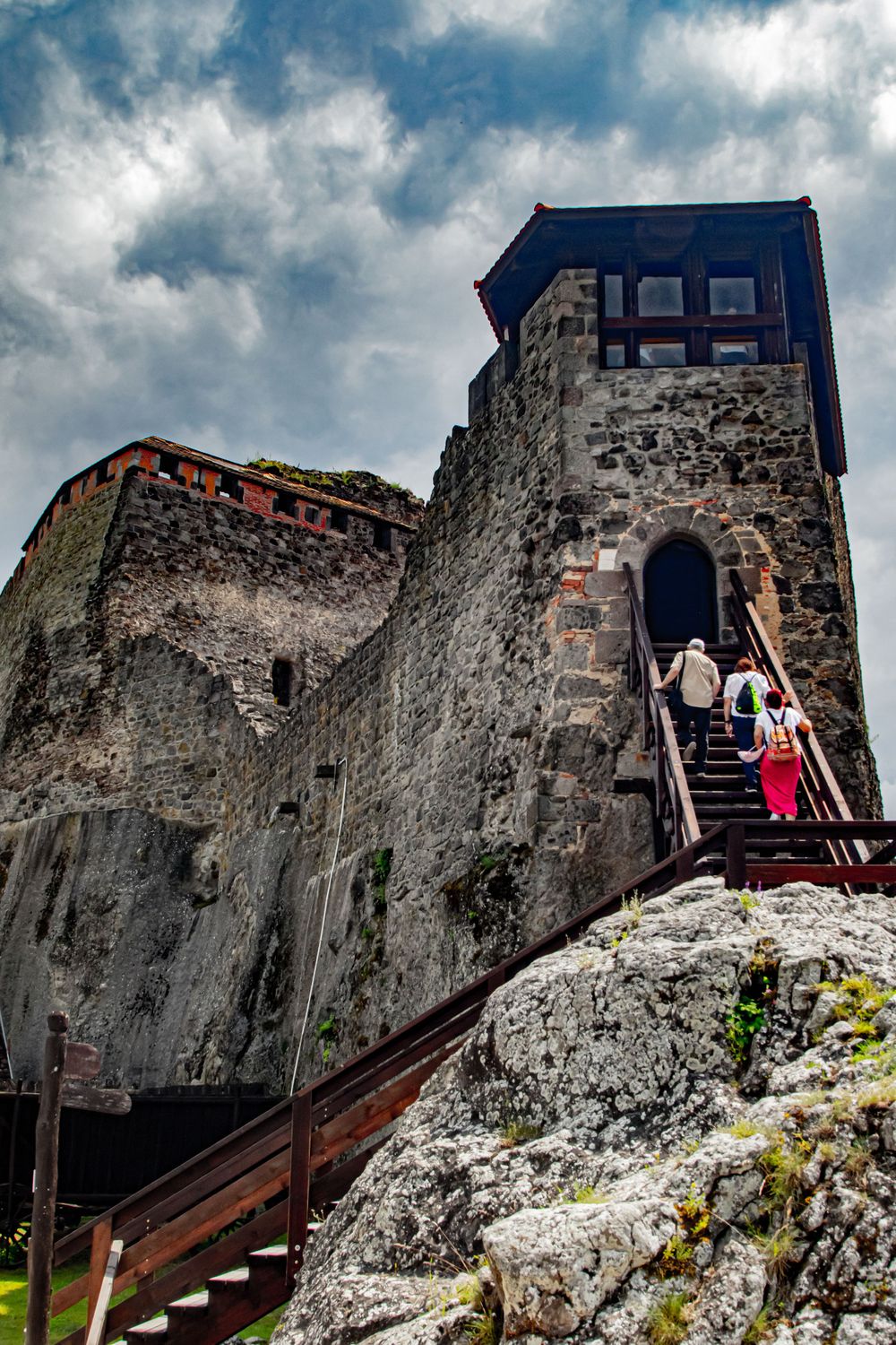 Medieval stone fortress tower with wooden staircase against dramatic cloudy sky.