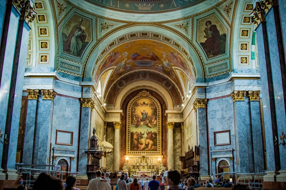 Ornate cathedral interior with blue-tinted marble columns and painted dome ceiling leading to an elegant gilded altar.