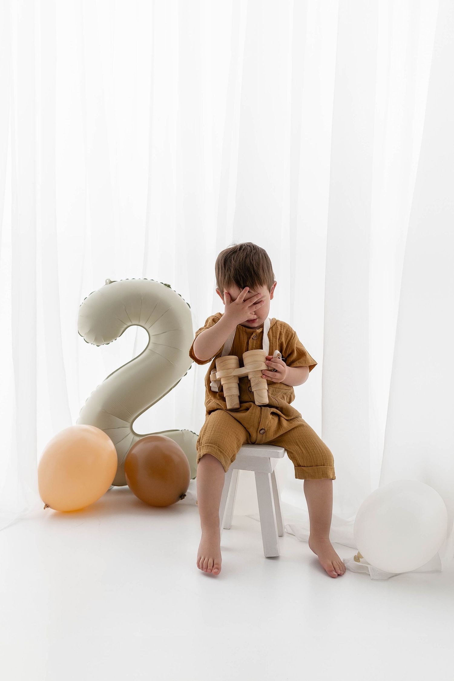 Child in brown overalls covers face while sitting on white chair with number 2 balloon and party decorations.