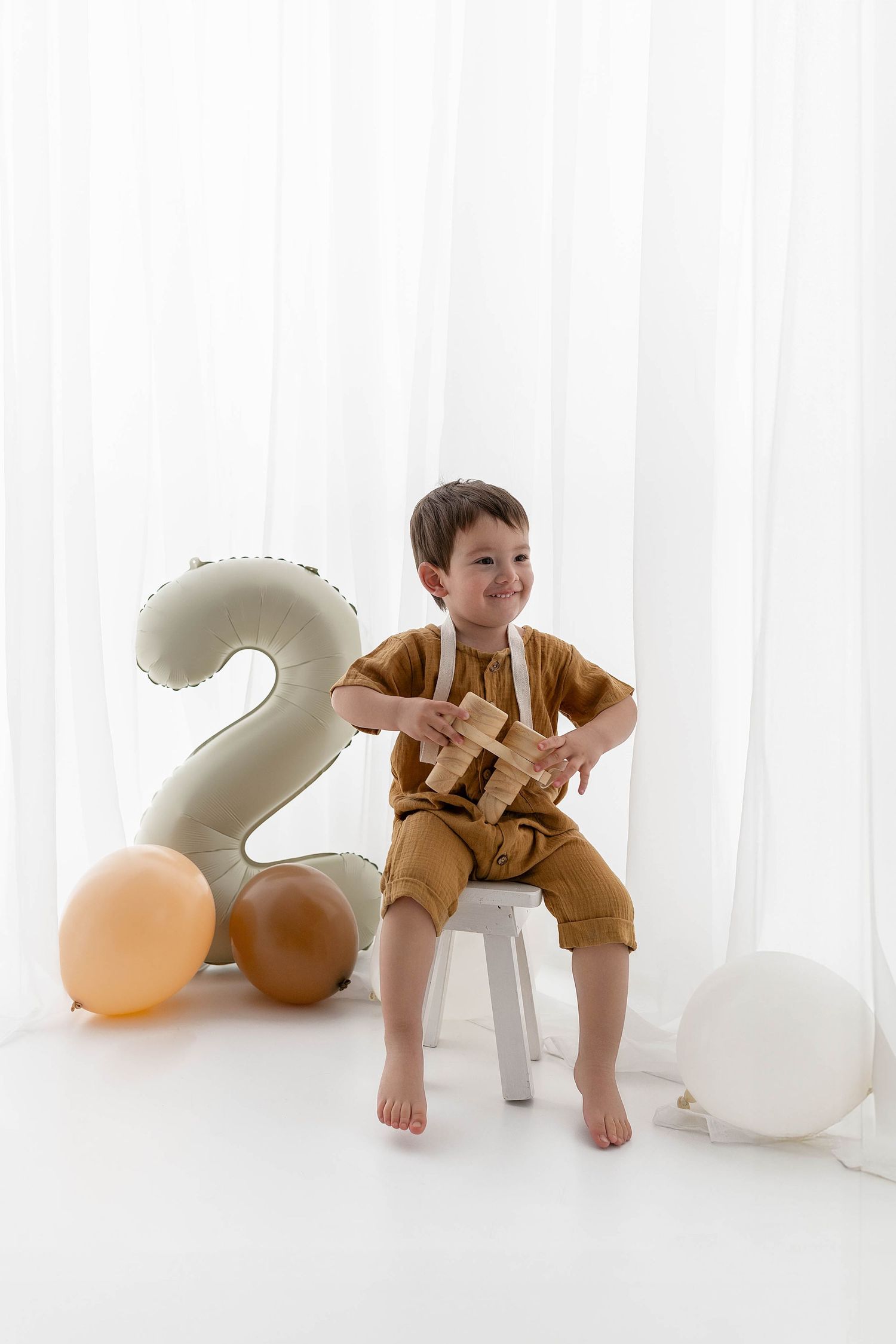 Child sits on white stool examining something while number 2 balloon and neutral party decor surround them.