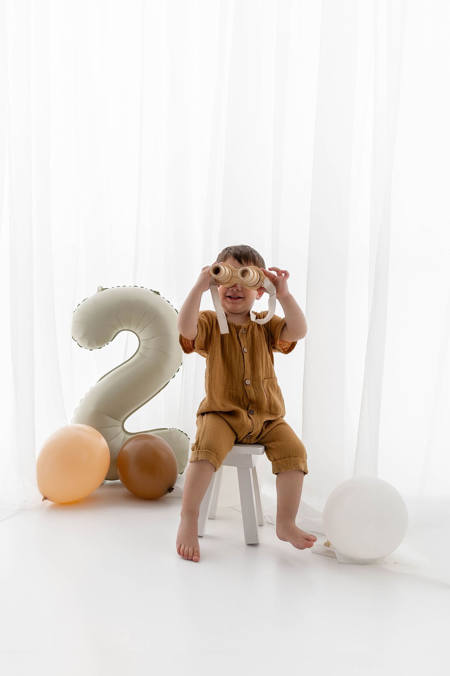 Toddler in brown romper plays expressively while sitting on white chair next to large number 2 balloon and party decor.