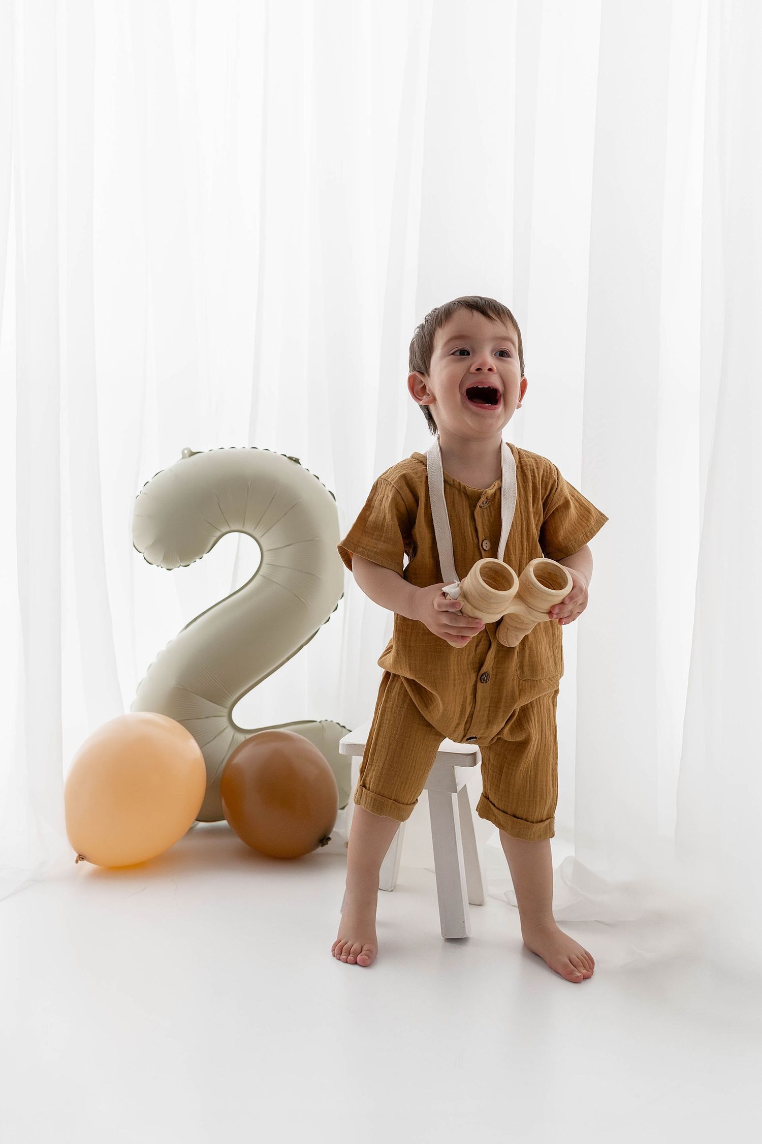 Joyful child in mustard outfit standing by number 2 balloon with toys.