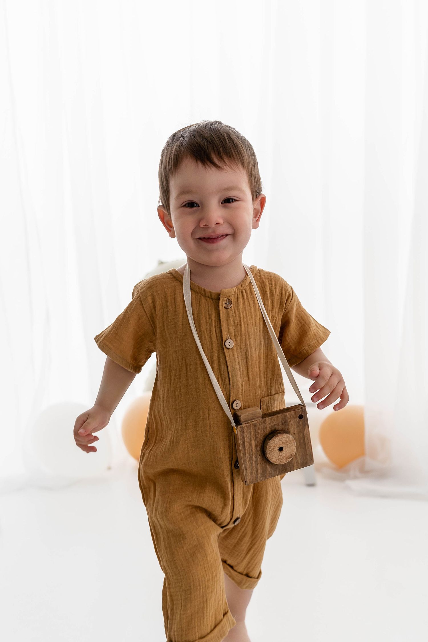 Child in mustard romper with toy camera smiling against white background.