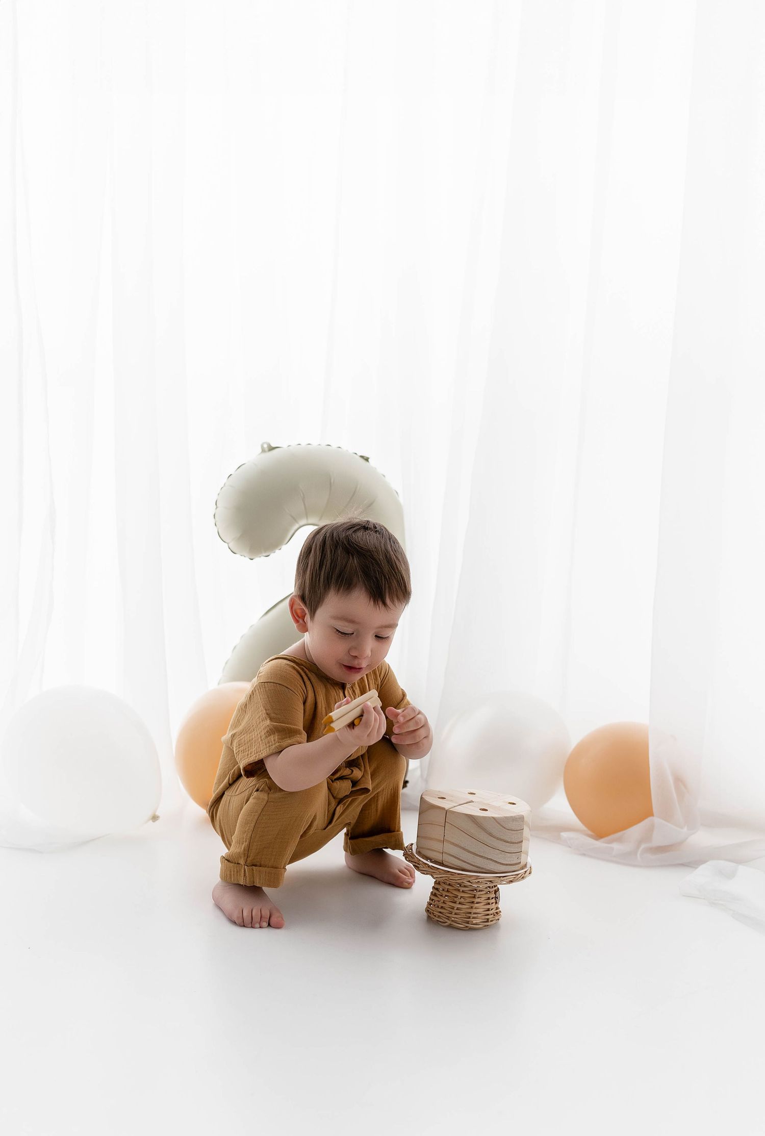 Small child in caramel outfit crouches down to examine a tiny birthday cake with neutral party decorations.