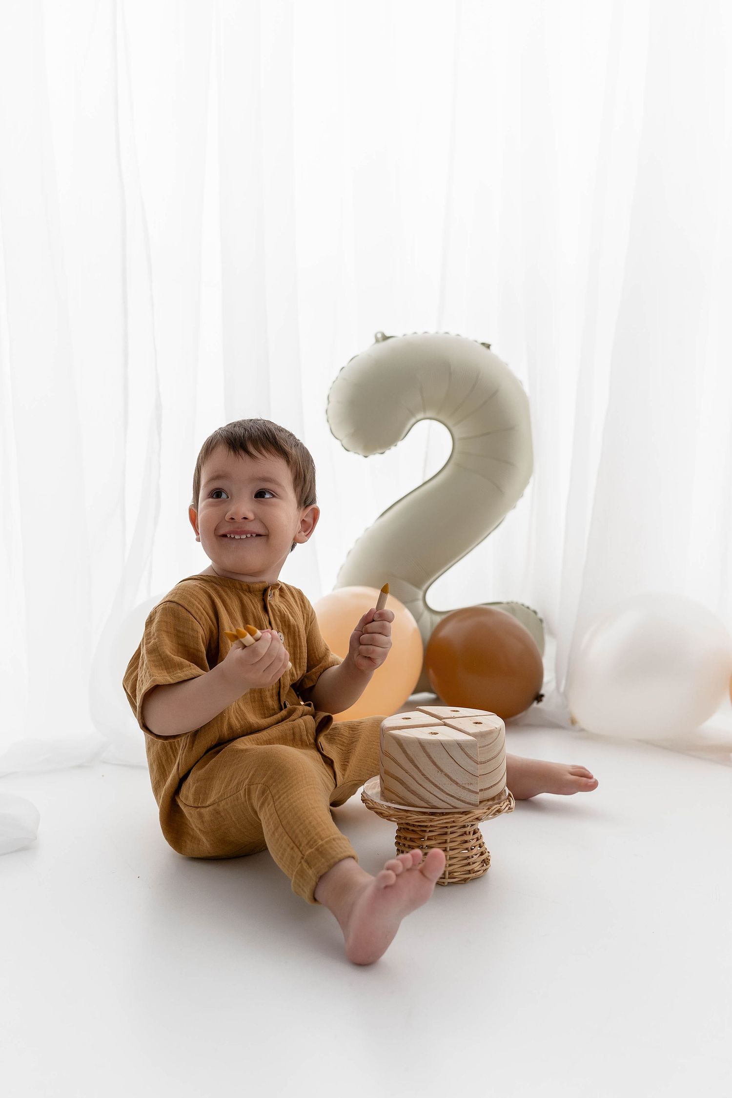 Child in mustard-colored outfit sits on white chair with number 2 balloon and birthday decorations.