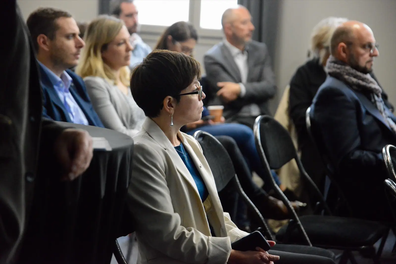 Femme en tailleur clair assise parmi d’autres personnes lors d’une conférence ou d’un colloque professionnel.
