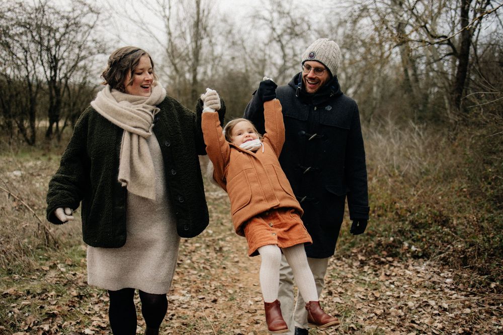 A family walks together on a leaf-covered path through a wooded area on an autumn day.