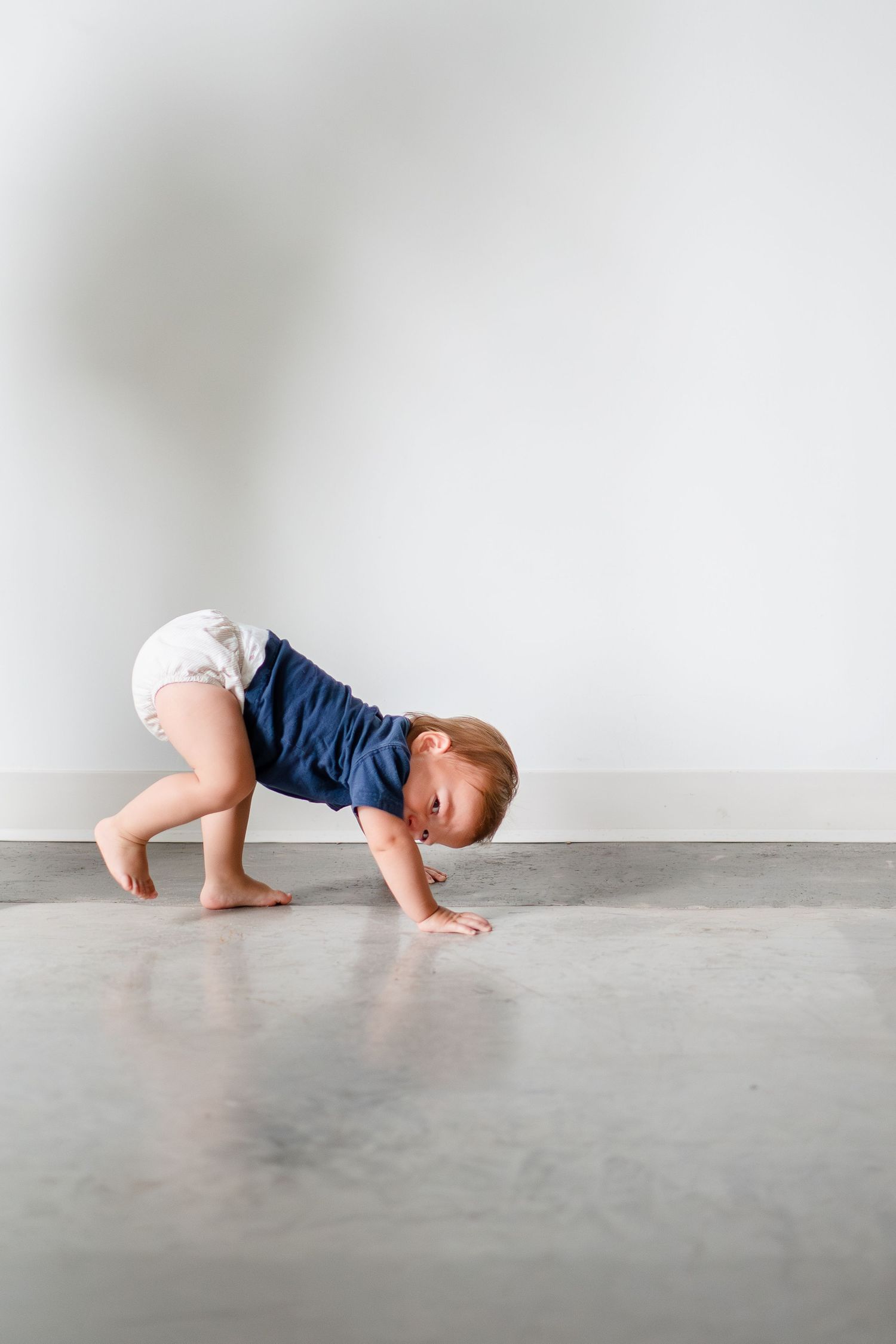 A toddler wearing blue shorts and a white diaper crawls on a grey concrete floor against a white wall.