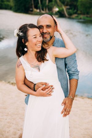 A loving couple embraces on a beach during a sunset photoshoot, with the bride wearing a white lace dress.