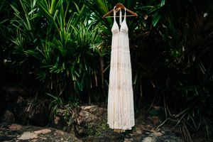 A white lace wedding dress hangs on a wooden hanger against lush tropical greenery background outdoors.