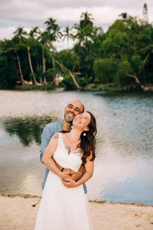 A couple embraces joyfully on a tropical beach with palm trees and calm waters in the background.