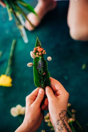 Tattooed hands arrange delicate flowers and greenery against a dark teal surface during a floral design session.