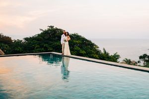 A couple in white stands at the edge of an infinity pool overlooking the ocean at sunset.