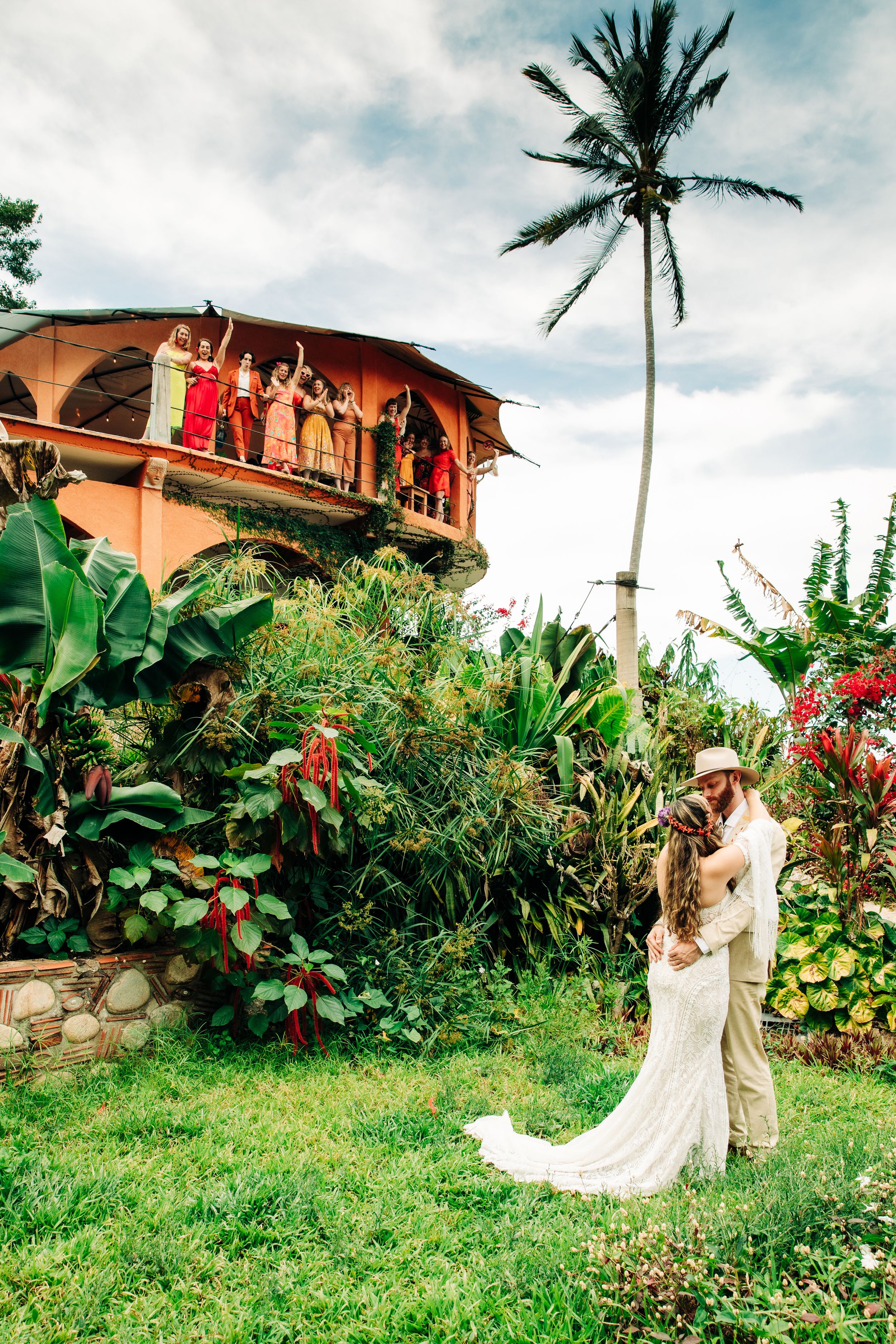 Tropical wedding celebration in Hawaii with wedding party watching from balcony above the couple in a lush garden setting.
