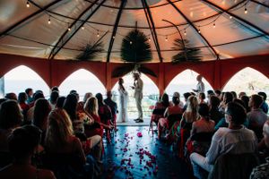 A wedding ceremony takes place under a red-draped gazebo overlooking the water as guests watch from their seats.