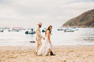 A couple walks hand in hand on a sandy beach with boats anchored in the bay and hills in the background.