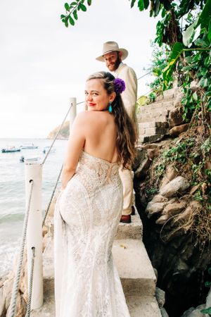 A couple in wedding attire stands on stone steps by the ocean, with lush greenery visible along a rocky path.