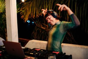 DJ in green striped shirt dancing and performing at night with laptop and equipment setup at outdoor venue.