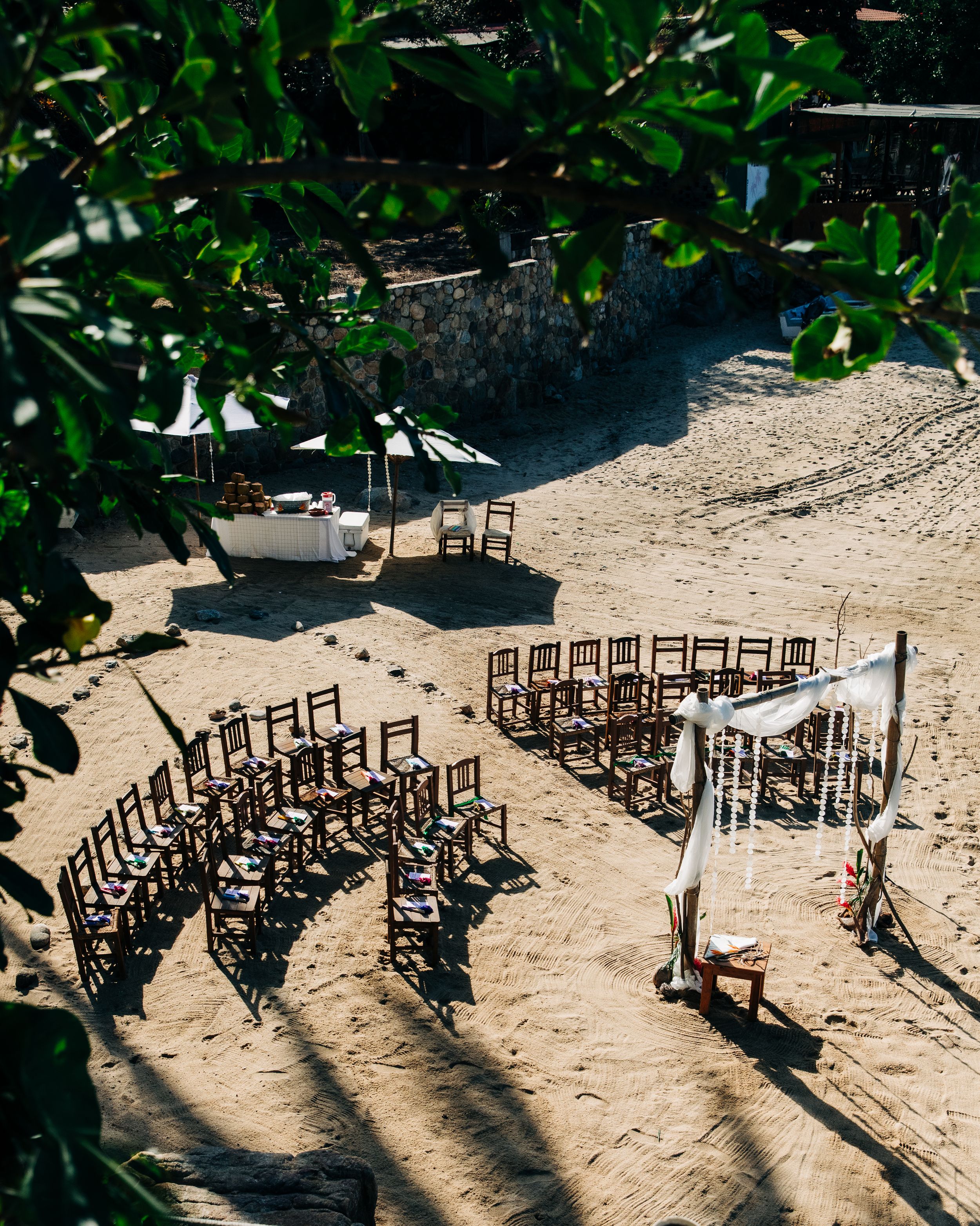 Curved rows of wooden chairs arranged for a beach wedding ceremony with white arch against tropical foliage backdrop.