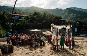 A beach wedding ceremony setup with guests gathered under a white arch canopy against a mountainous tropical backdrop.