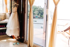 White wedding dress hangs by sliding glass doors with sheer curtains overlooking a balcony view.