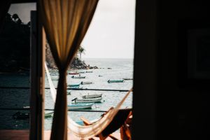 View from inside room through curtains showing tropical beach with moored boats and someone relaxing in hammock.